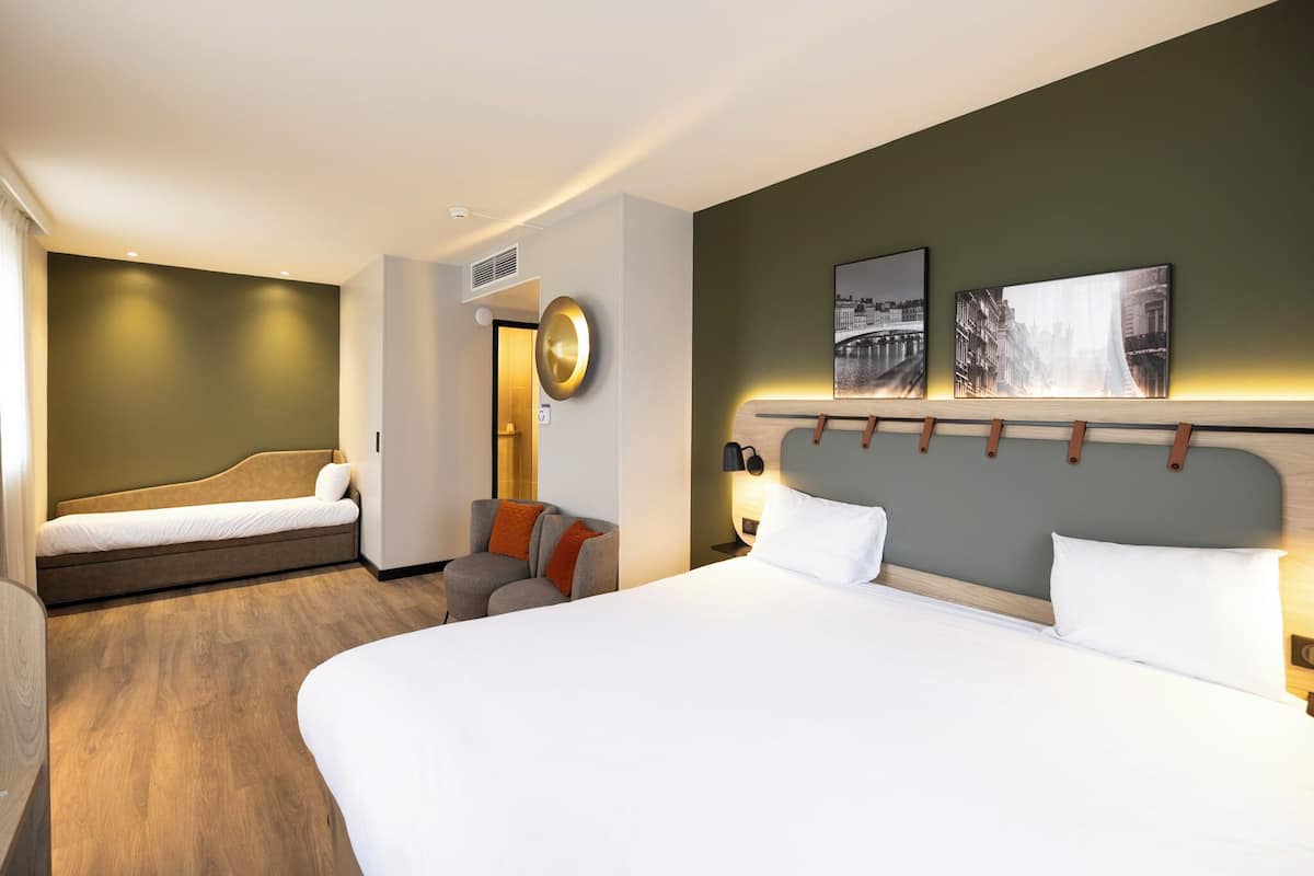 A hotel room with a large bed, a smaller sofa bed, a chair, and a wall-mounted lamp.