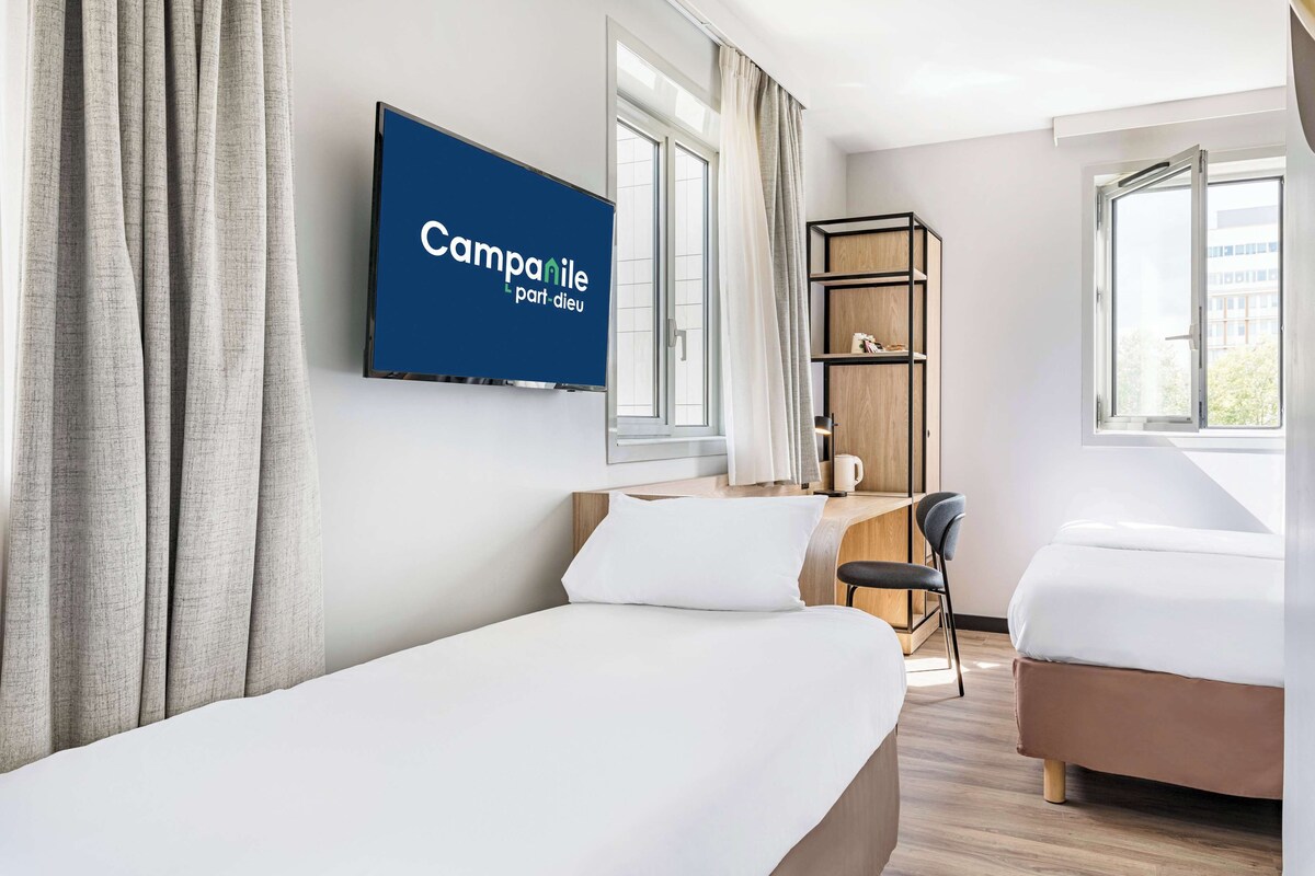 Habitación de hotel con dos camas, un escritorio con silla, un televisor montado en la pared y una estantería.