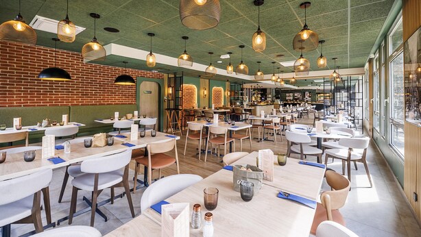 A spacious restaurant with multiple dining tables, hanging pendant lights, and large windows providing natural light.