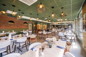 Un restaurante espacioso con varias mesas, lámparas colgantes y grandes ventanales que dejan entrar luz natural.