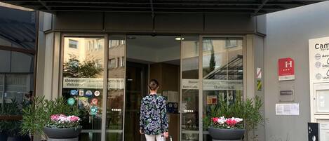 A hotel entrance with a glass door, potted plants, and a sign indicating room availability.