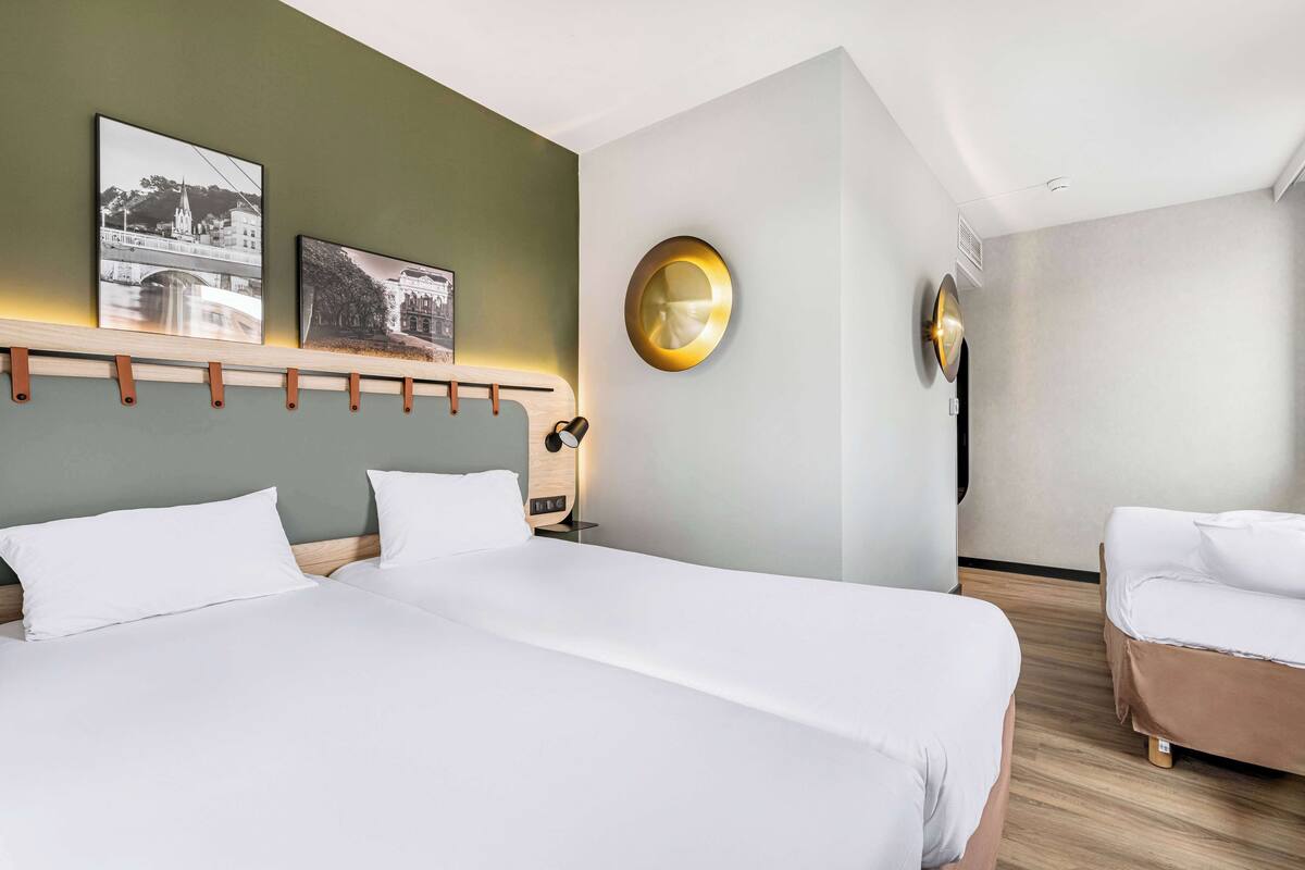 A hotel room with a large bed, a bedside table with a lamp, a round mirror, and two framed pictures on the wall.
