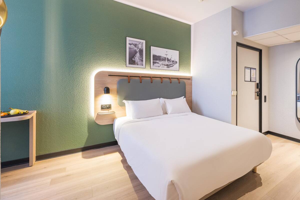A modern hotel room with a bed, a nightstand, a mirror, and a green wall with two framed pictures.