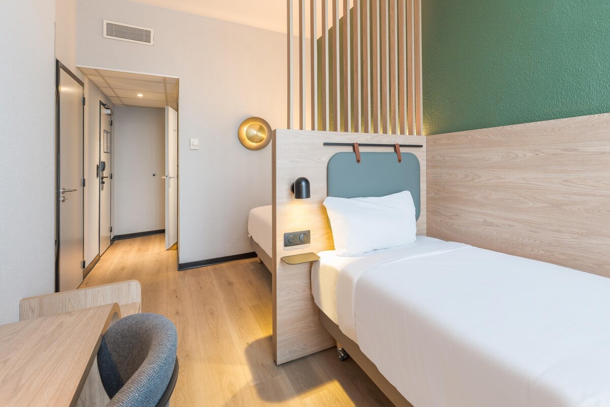 Habitación de hotel pequeña con una cama individual, un escritorio y una silla. Se ve una puerta que conduce a otra habitación y una lámpara fijada a la pared.