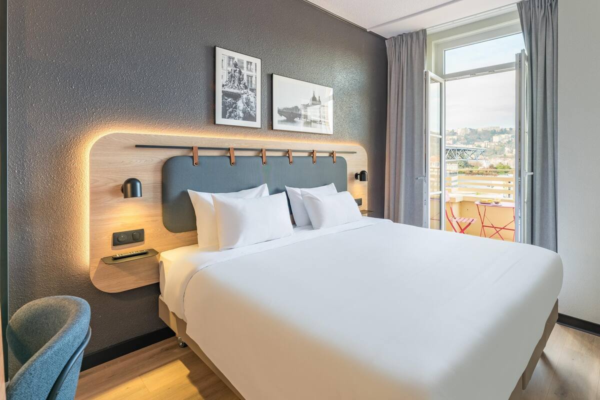 A modern hotel room with a large bed, a headboard with built-in lighting, a nightstand, a desk, and a view of the city through a large window.