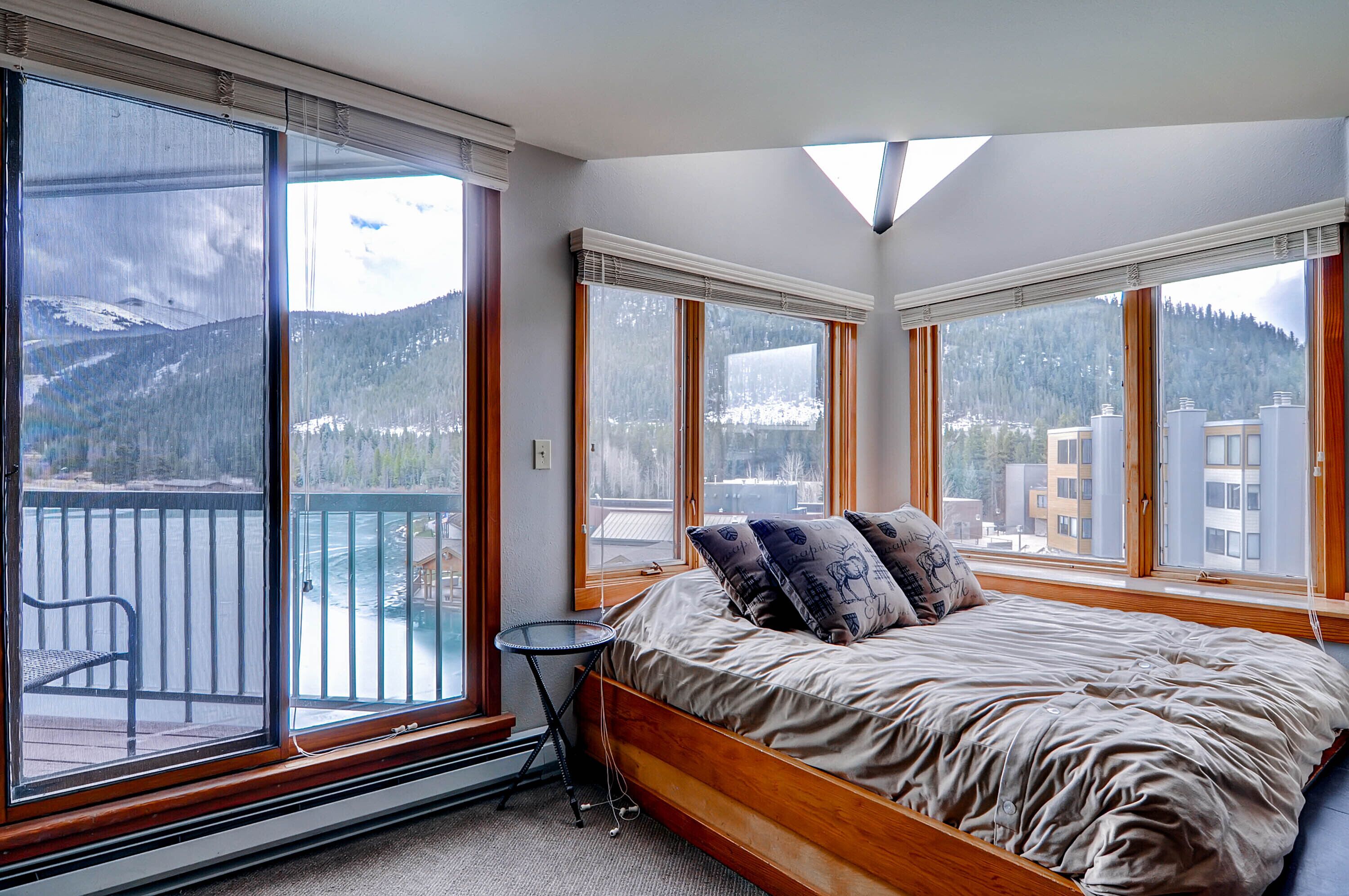 A bedroom with a large bed, a small table, and a view of mountains and buildings through large windows.