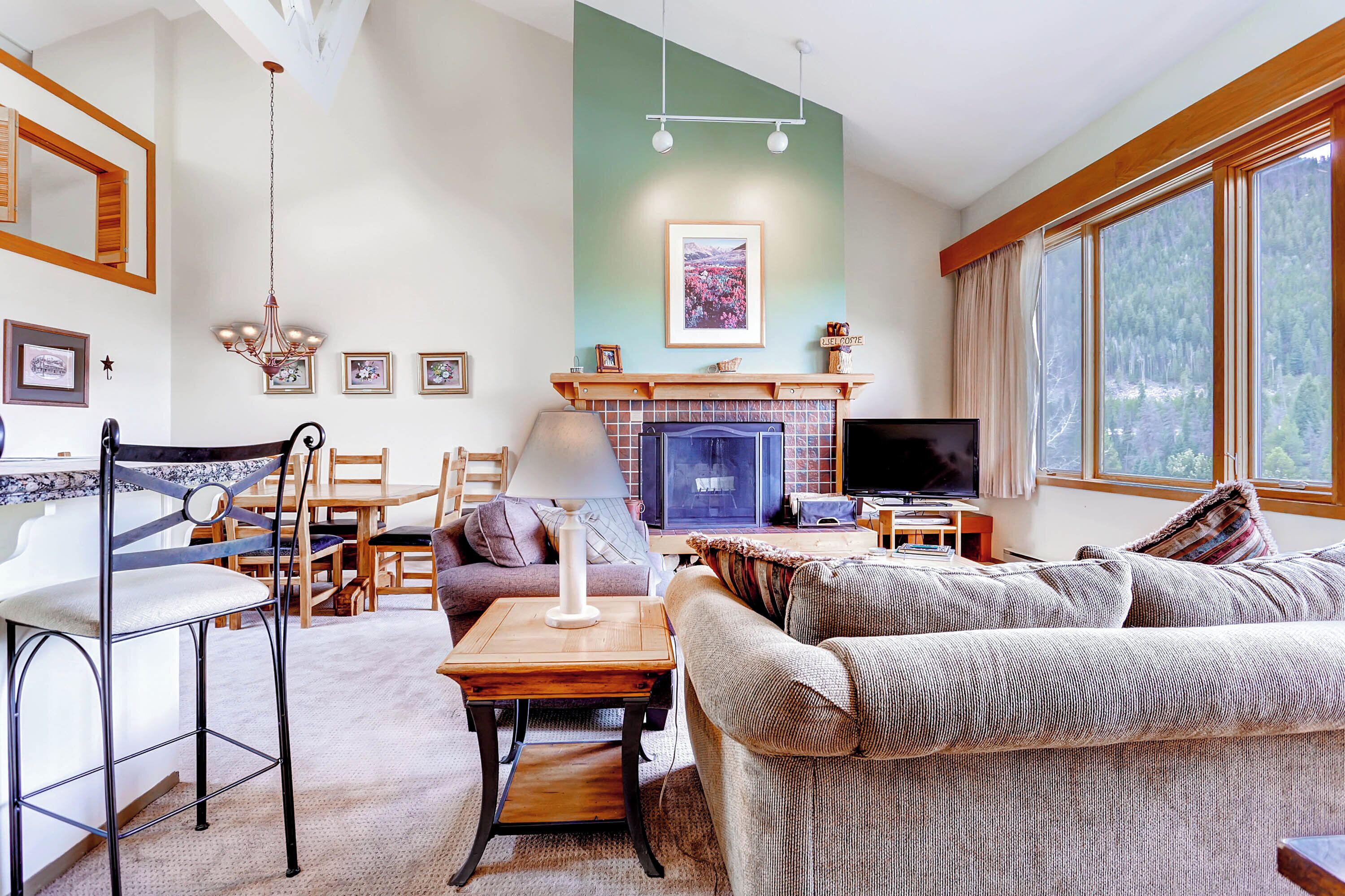 A cozy living room with a fireplace, a television, and a large window with a view of trees.