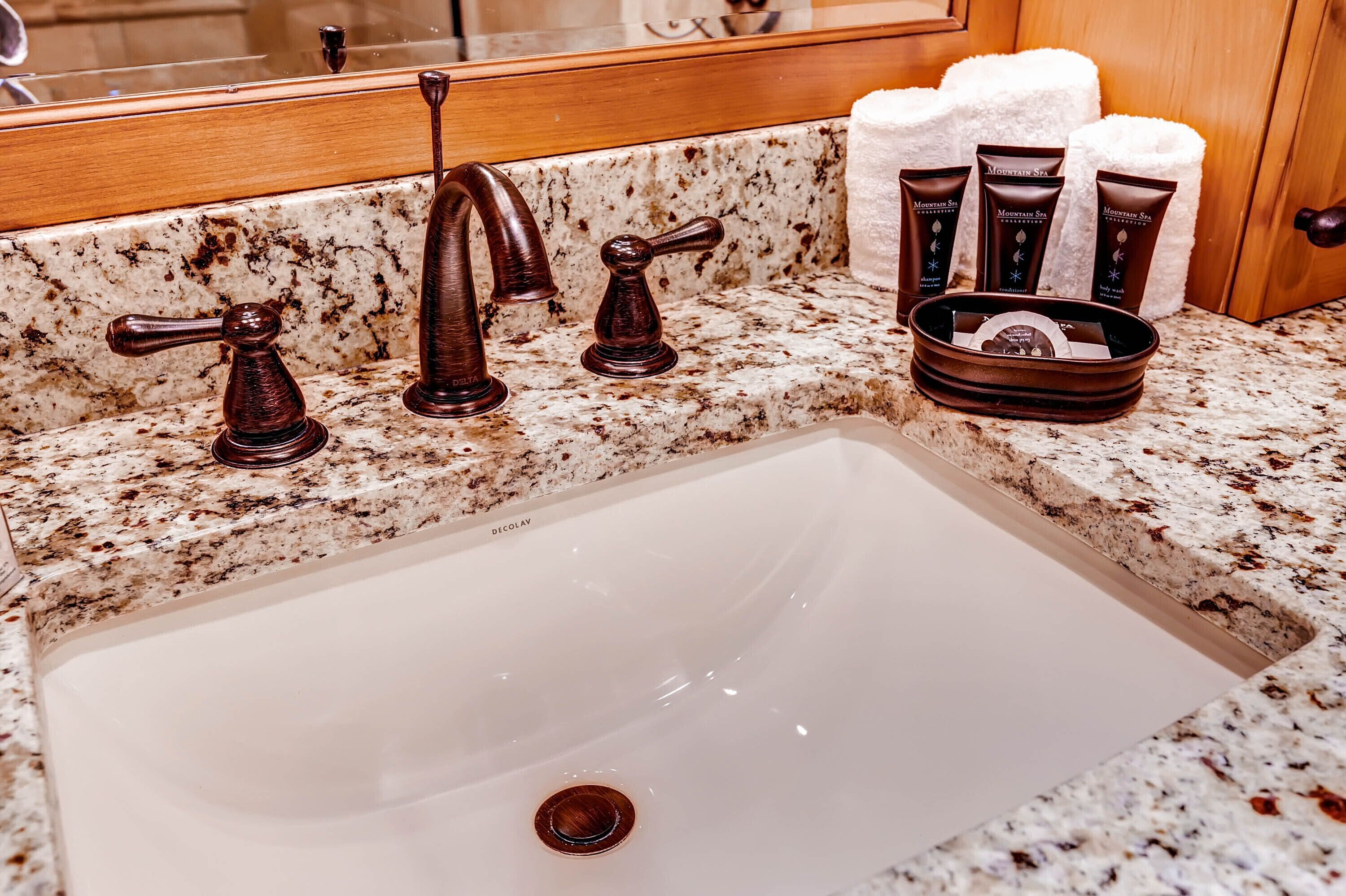 A bathroom with a granite countertop, a white sink, and three dark-colored faucets. There are three bottles of Mountain Spa body wash and a basket of white towels.