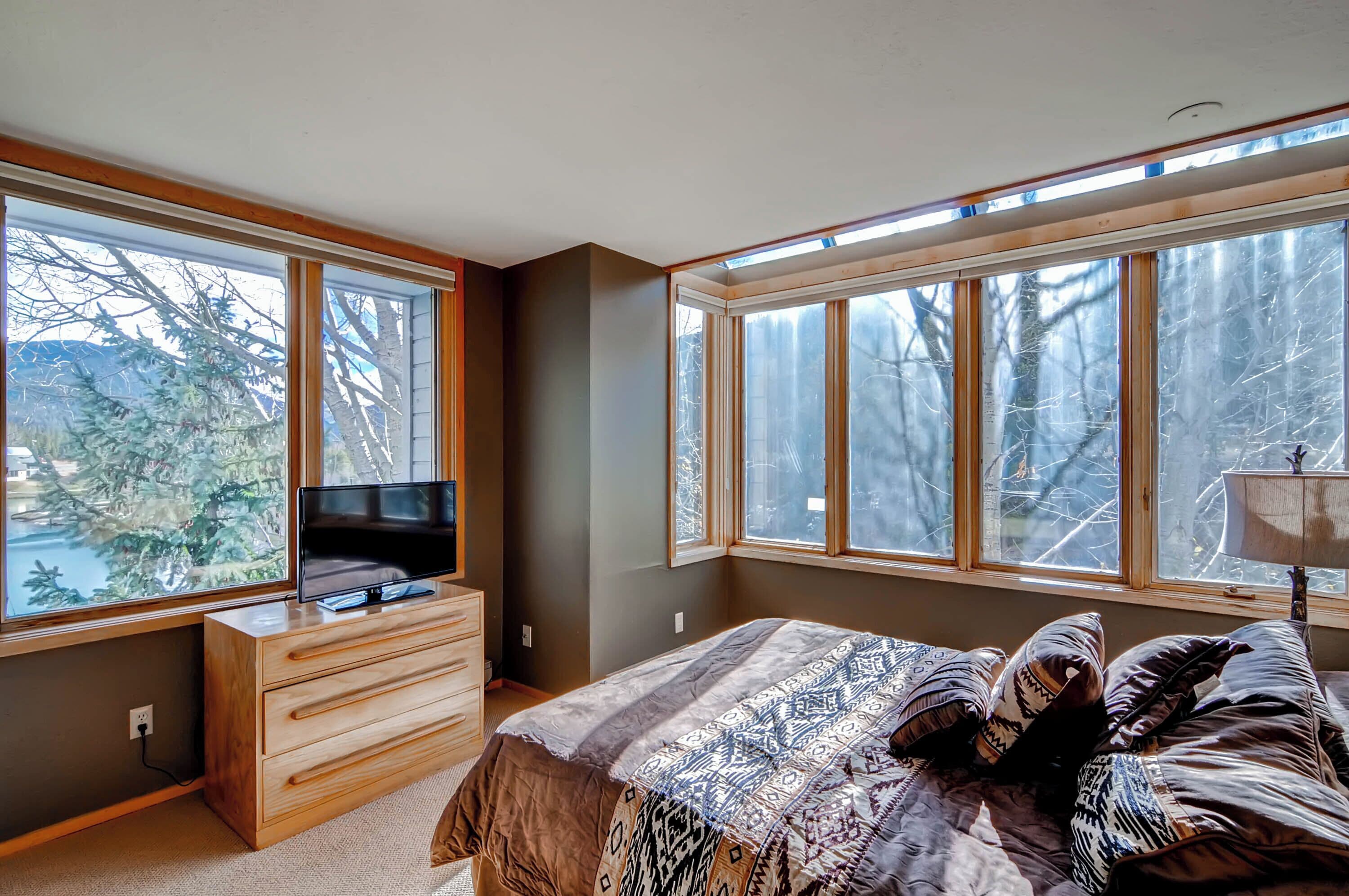 Ein Schlafzimmer mit einem Bett, einem Fernseher auf einer Holzkommode und großen Fenstern mit Blick auf Bäume.