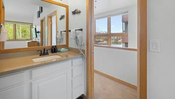 A bathroom with a large mirror, a sink, and a shower area. There is a window with a view of buildings and trees.