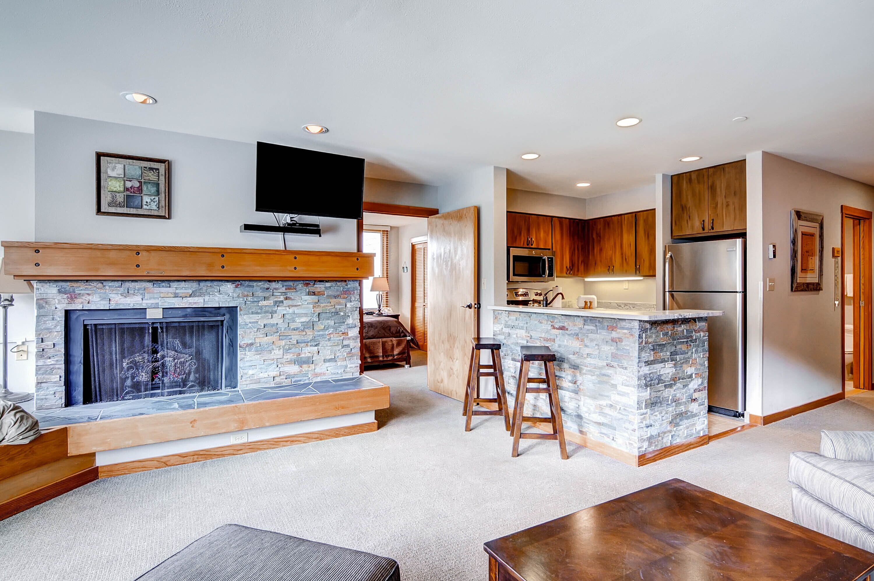 A modern living room with a stone fireplace, a flat-screen TV, and a kitchen area with wooden cabinets and stainless steel appliances.