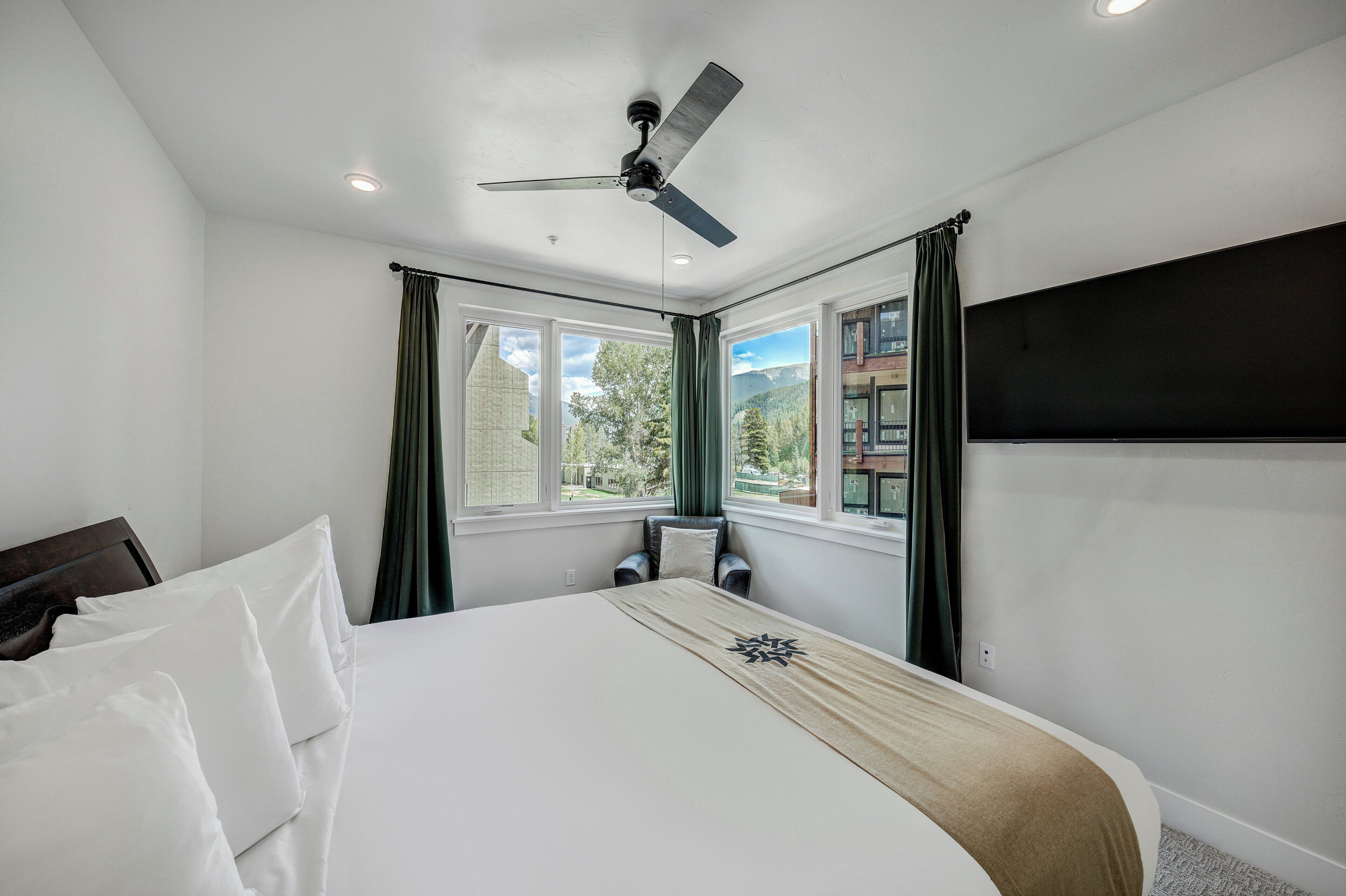 A hotel room with a bed, a ceiling fan, large windows, and a view of the outdoors.
