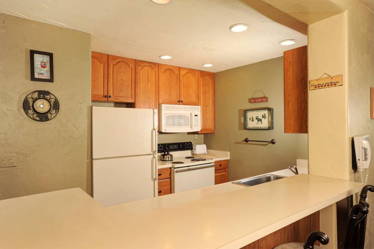 A compact kitchen with wooden cabinets, a white refrigerator, a microwave, and a stove. There is a clock on the wall and a sign that says "Eat-Sleep-Ski".