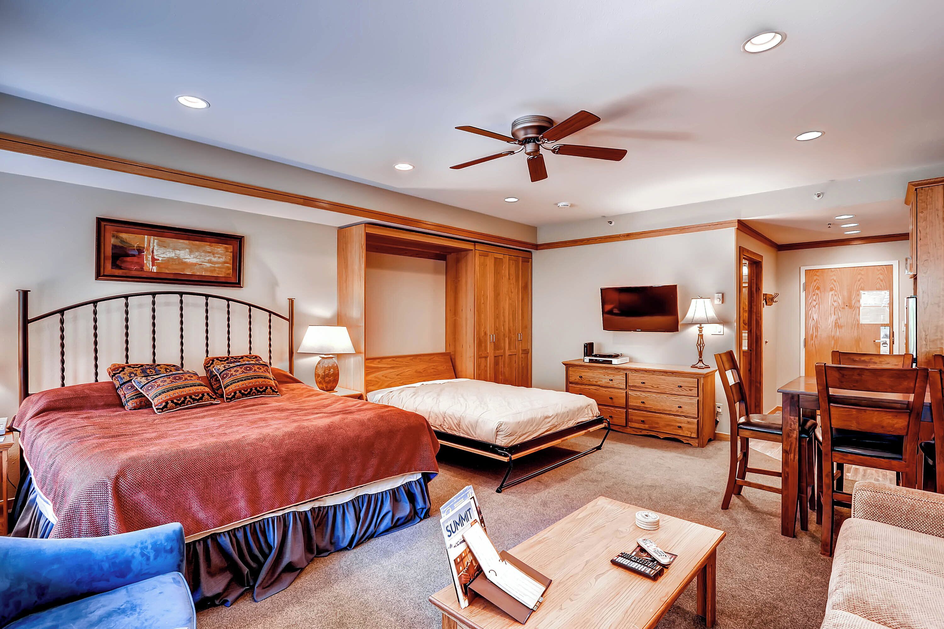 A hotel room with a large bed, a desk, a chair, a ceiling fan, and a TV.