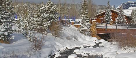 A snow-covered landscape with a wooden bridge, a building with a sloped roof, and a river partially frozen with snow.