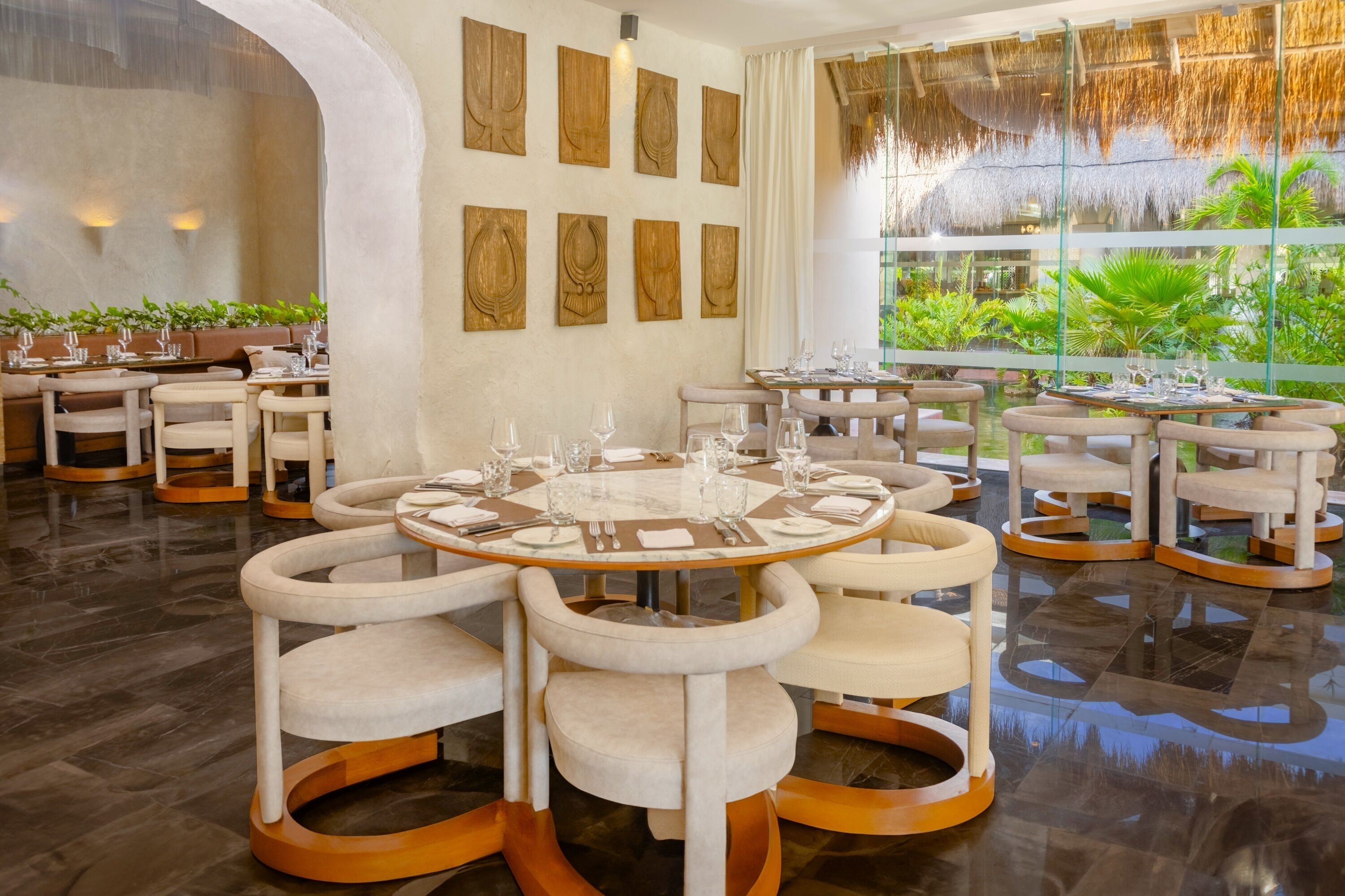 A modern dining area with round tables and cushioned chairs, surrounded by a glass wall with a view of greenery.