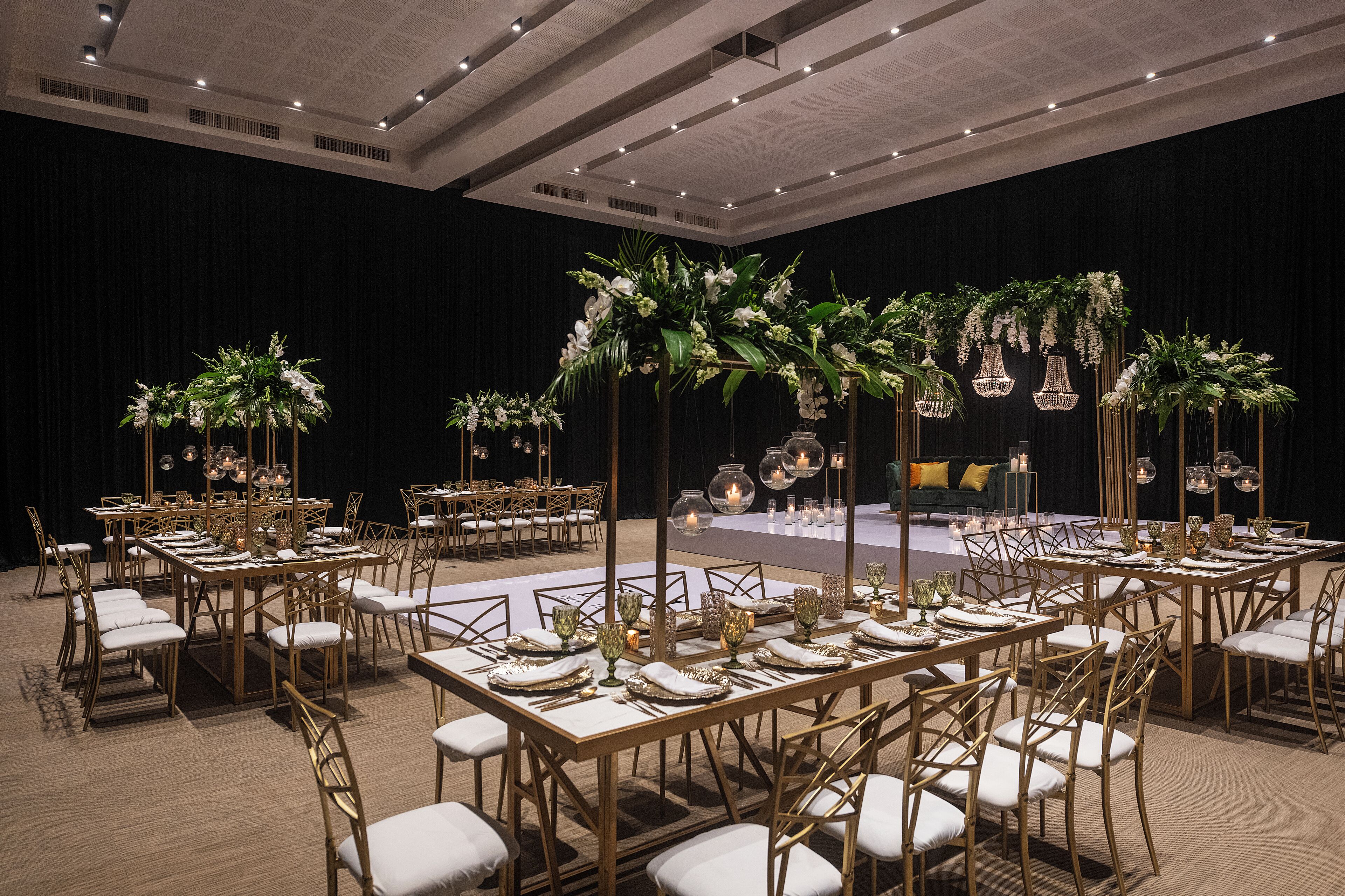 A banquet hall with long tables set for dining, surrounded by chairs, and decorated with floral arrangements and hanging lights.