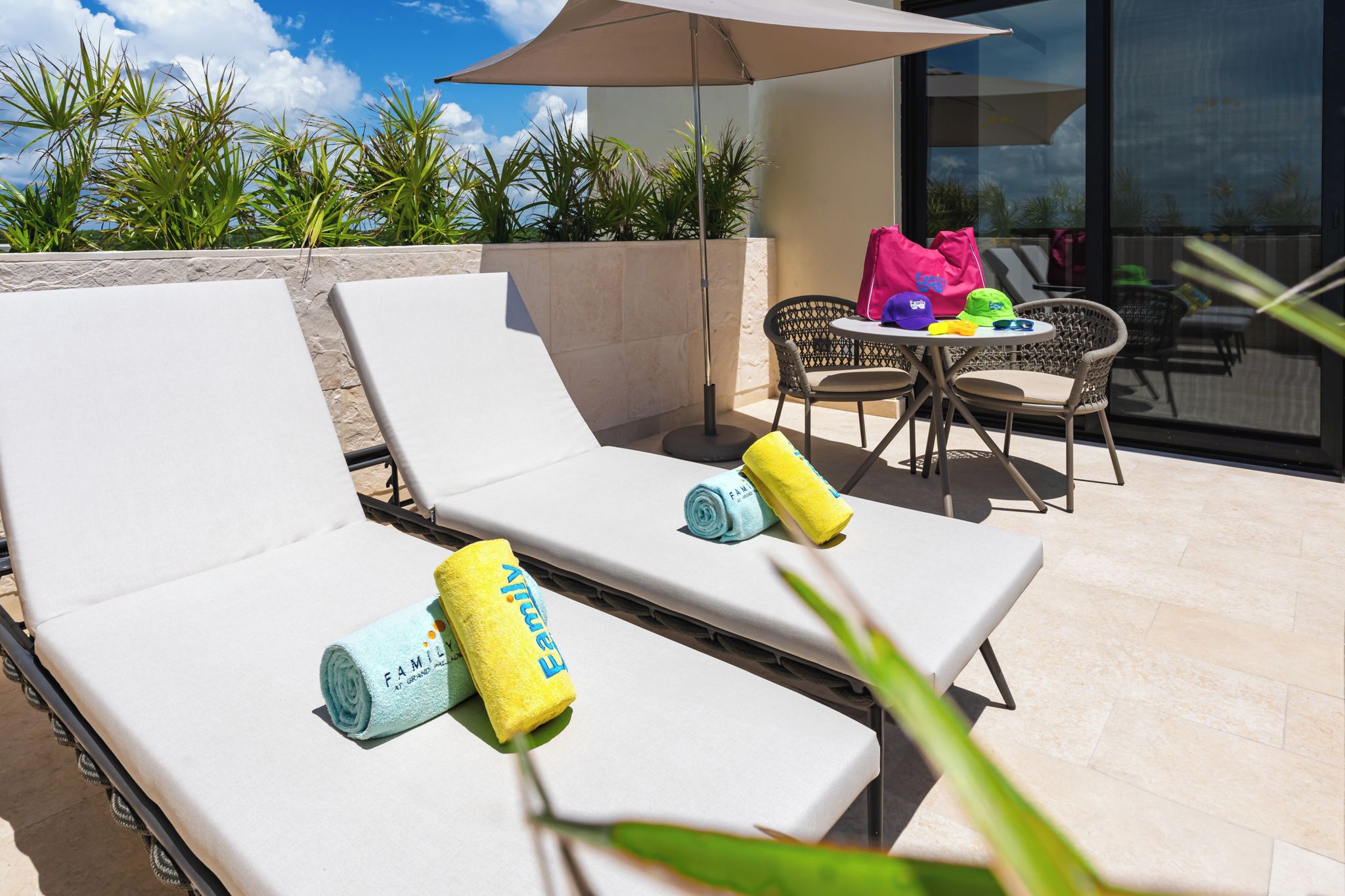 A sunlit outdoor lounge area with two white lounge chairs, a table with chairs, and a large umbrella providing shade.