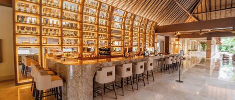 Zona de bar con techo alto y una pared de estantes llenos de botellas. Hay varios taburetes de bar y una barra.