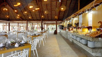 Un comedor amplio con sillas y mesas blancas, una barra de buffet con diversas opciones de comida, y un techo de paja.