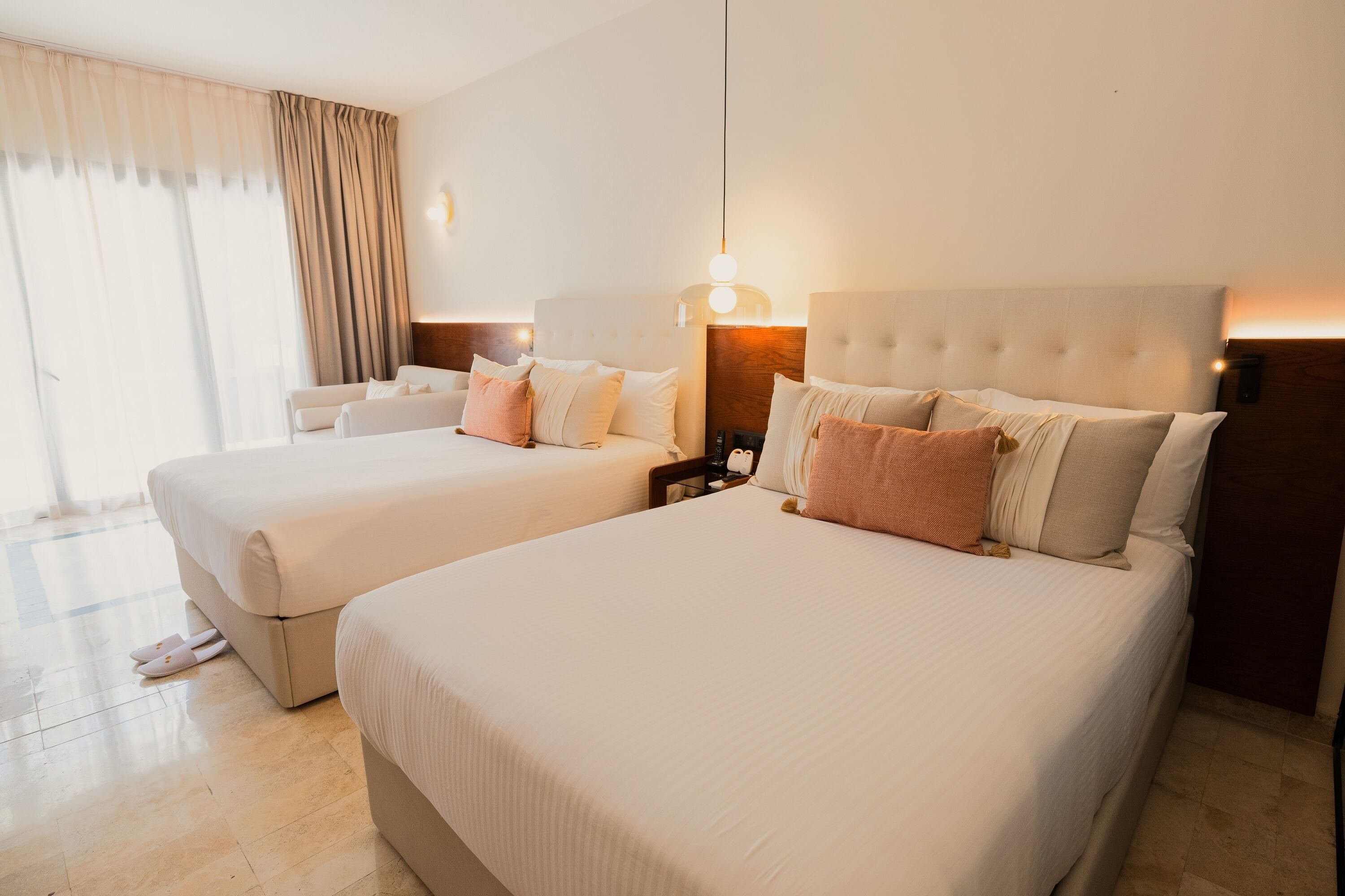 A hotel room with two beds, a large window with curtains, and a nightstand with a lamp.