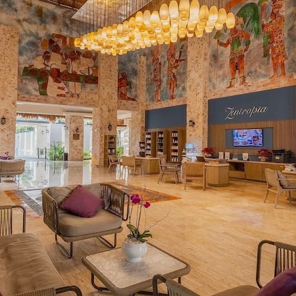 A spacious lobby with a high ceiling, a central seating area with sofas and chairs, a reception desk, and a large mural on the walls.