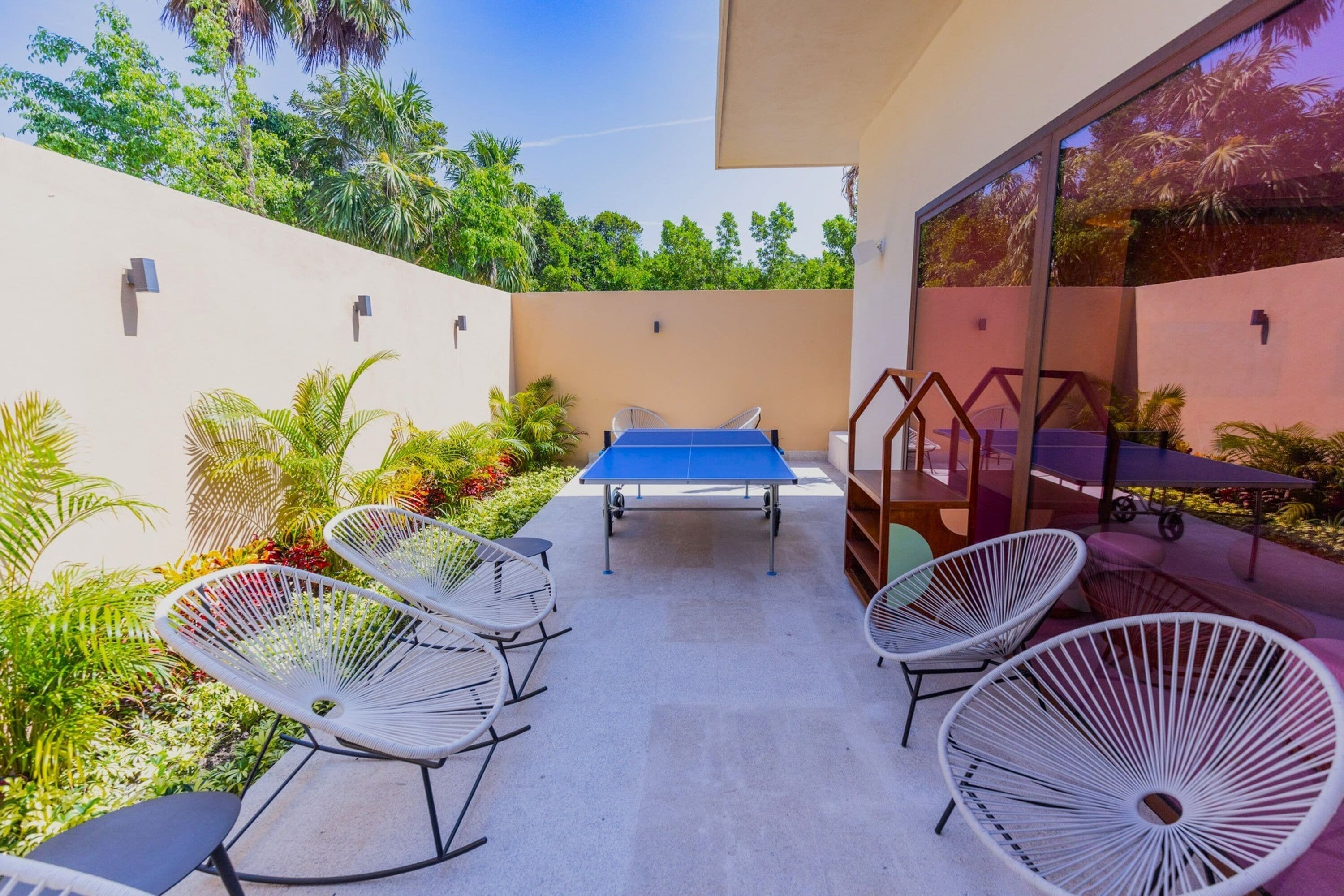 Un patio al aire libre con mesa de ping pong, sillas de alambre blancas y una zona de comedor con mesa y sillas.