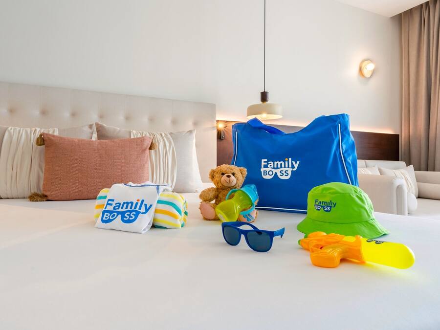 Quarto de hotel com cama, travesseiros, um urso de pelúcia, uma bolsa azul, um boné verde e uma prancha de snowboard amarela.