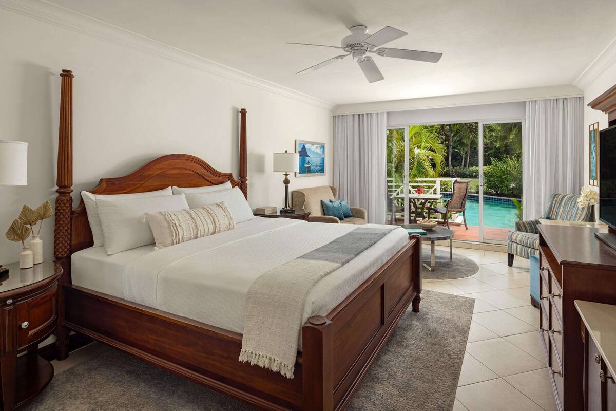 Ein geräumiges Schlafzimmer mit einem großen Bett, einem Deckenventilator, einem Nachttisch, einer Lampe und Blick auf einen Pool und eine Außenlounge.