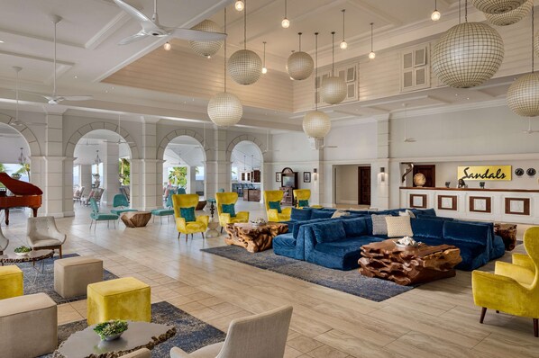 A spacious lobby with a grand piano, a blue sectional sofa, yellow armchairs, and a large wooden coffee table. - Sandals Grande St. Lucian - ALL INCLUSIVE Adults Only (Gros Islet)