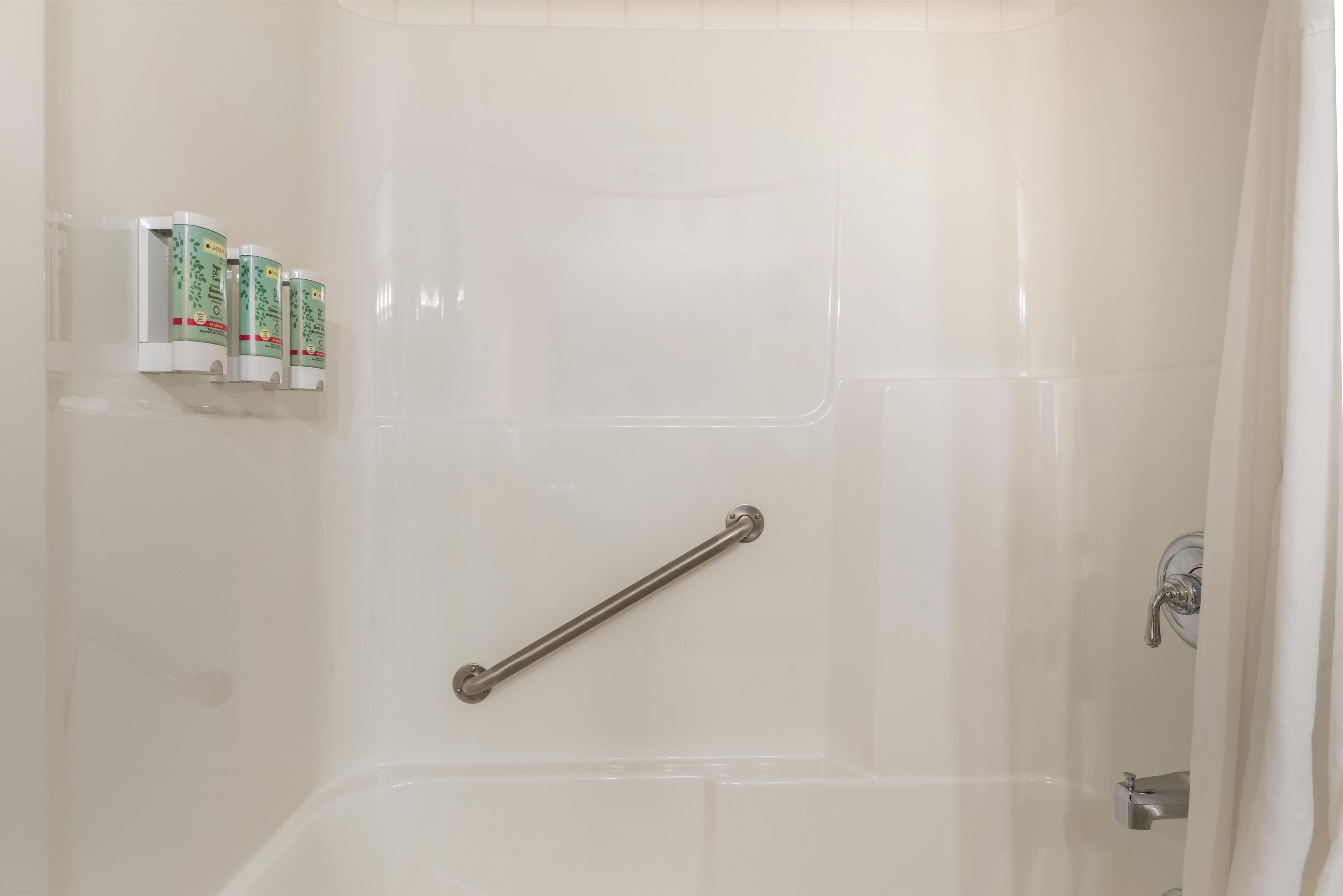 A bathroom with a white bathtub, a grab bar, and three bottles of shampoo on the wall.