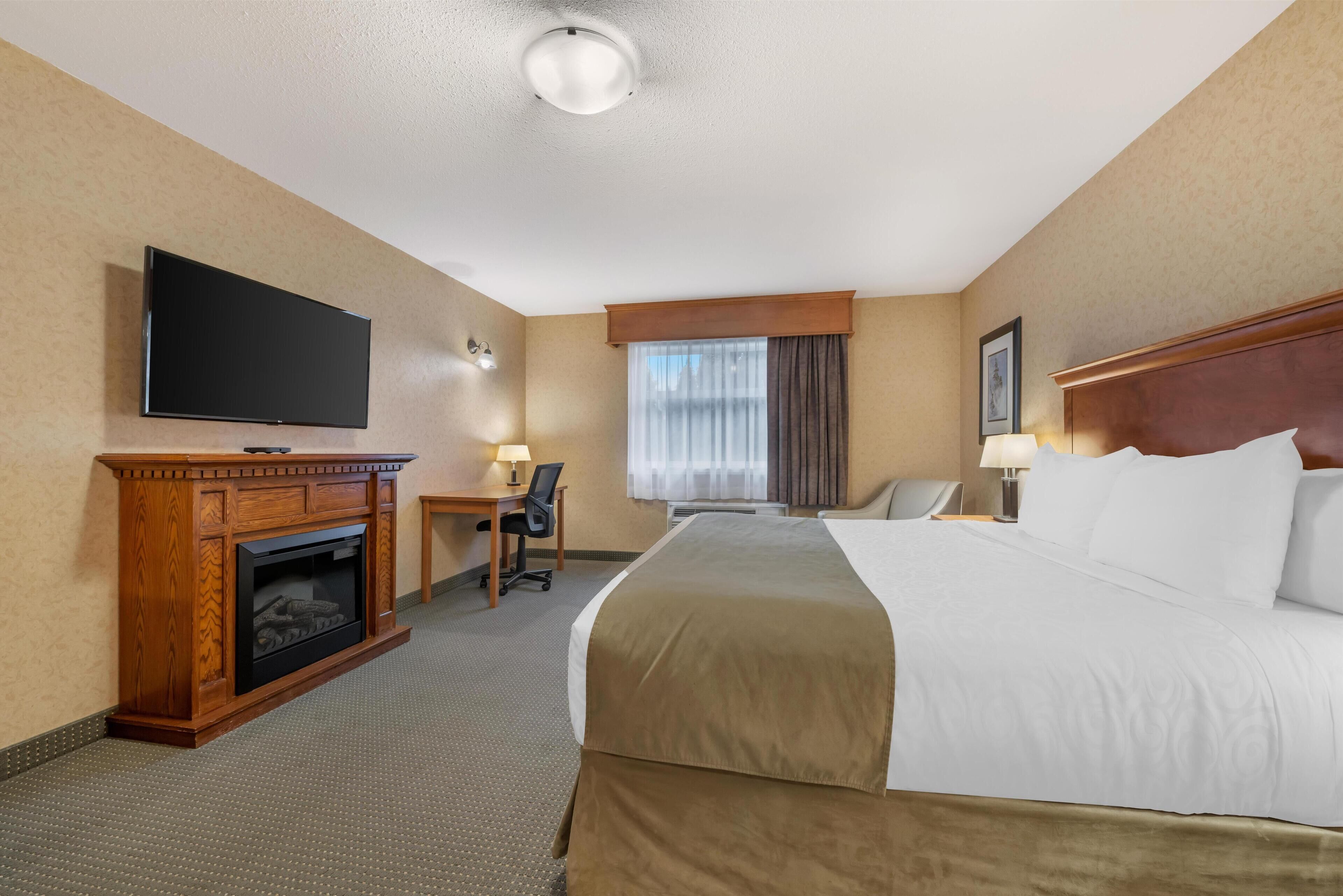 A hotel room with a bed, a television, a desk with a chair, a fireplace, and a window with curtains.
