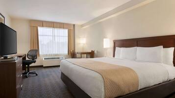 A hotel room with a large bed, a desk with a TV, a chair, a window with curtains, and a bedside table.