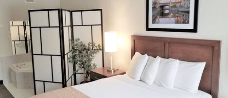 Habitación de hotel con cama, mesita de noche con lámpara, espejo, planta y un cuadro en la pared.
