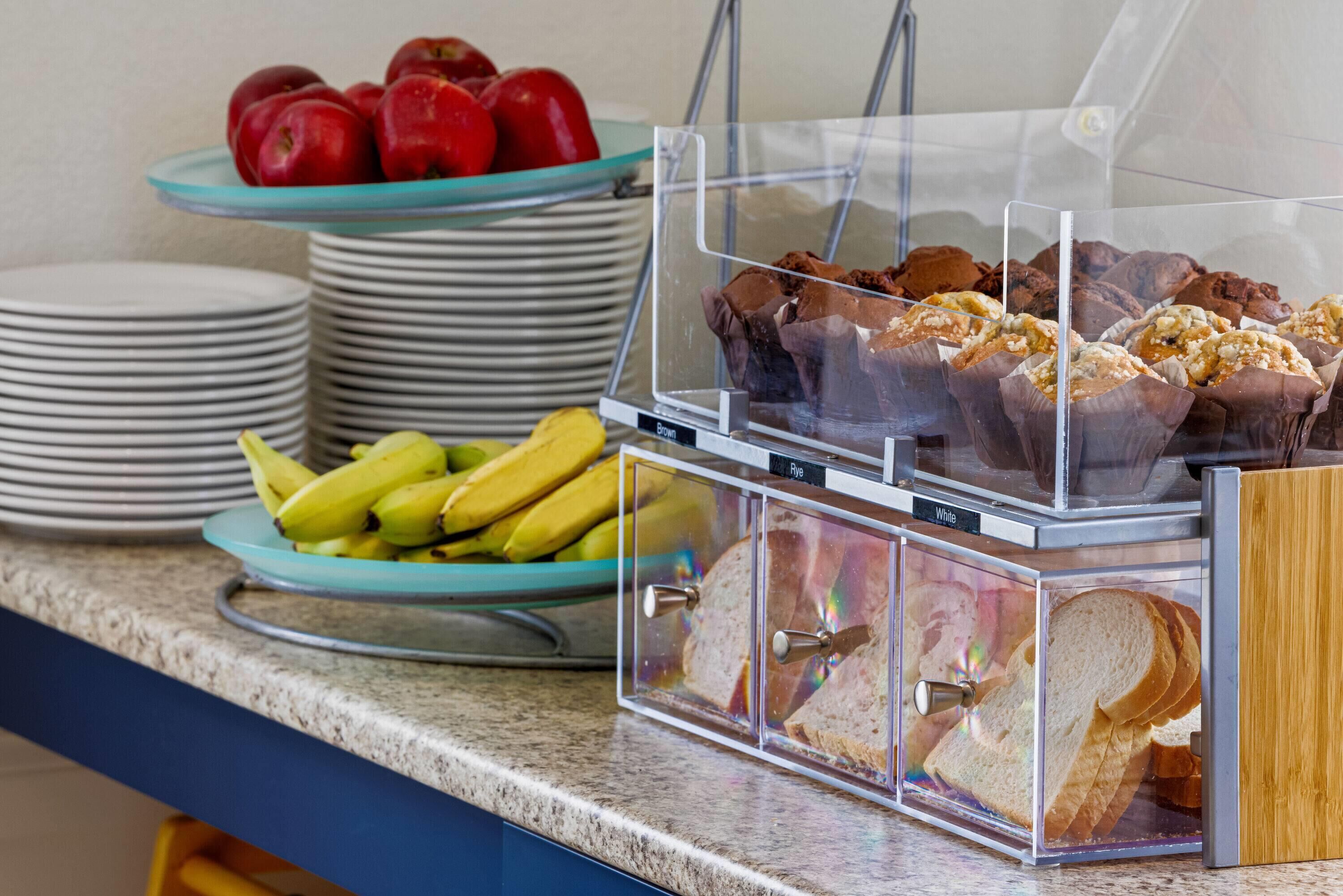 A buffet counter with clear glass containers holding sandwiches, muffins, and bananas.