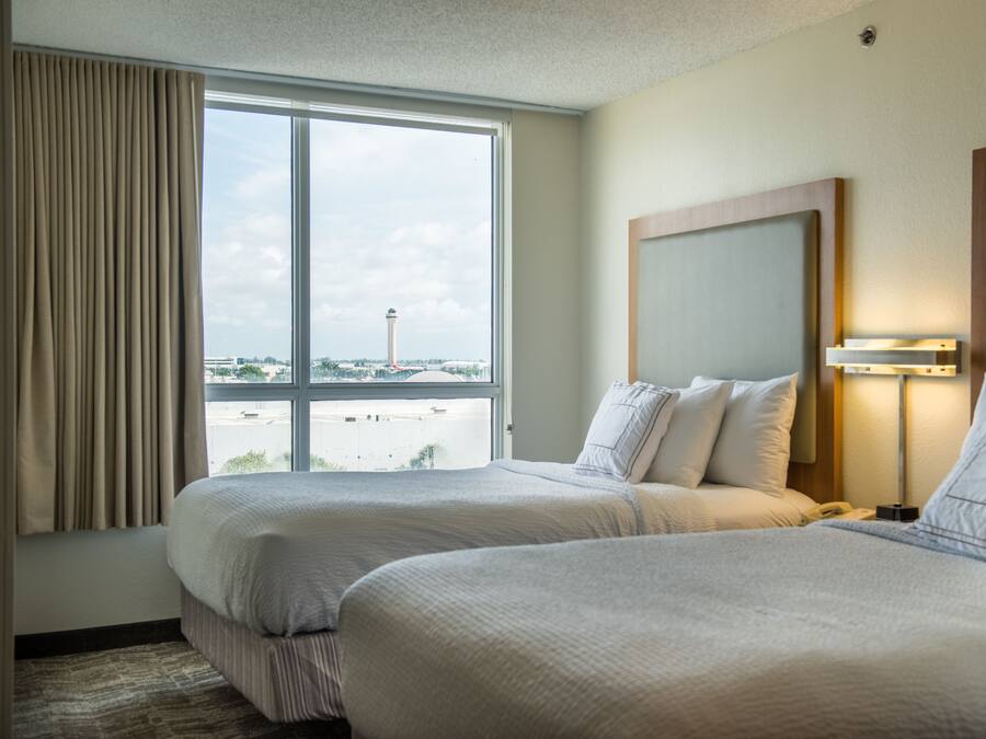 A hotel room with two beds, a large window with curtains, a view of the airport, and a nightstand with a lamp.