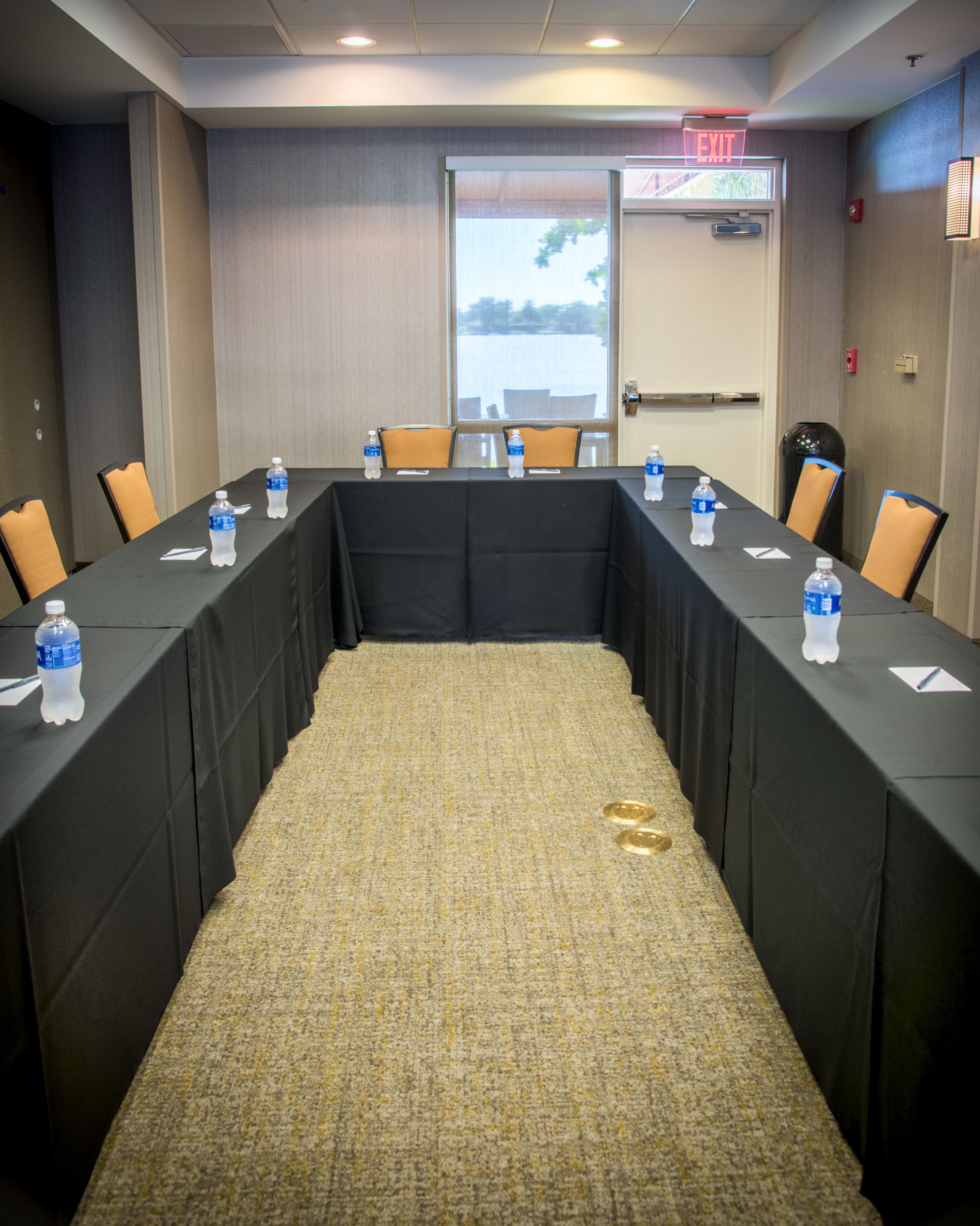 Un salón de conferencias con una larga mesa, sillas, botellas de agua y piso alfombrado.