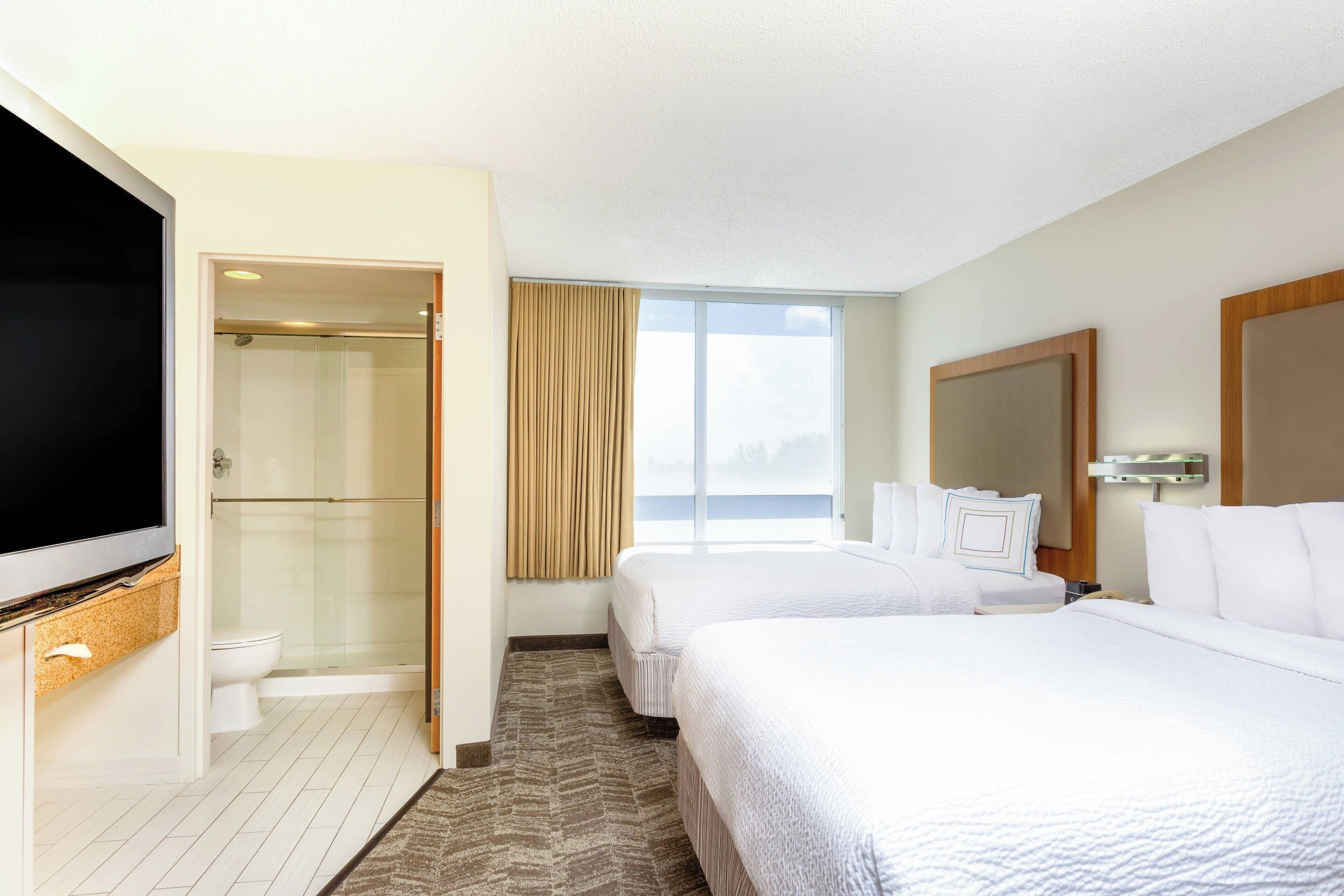 A hotel room with two beds, a TV, a shower, and a large window.