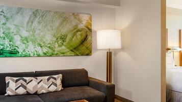 Habitación de hotel con una cama, un sofá con un cojín estampado, una lámpara de pie y un cuadro abstracto grande en la pared.