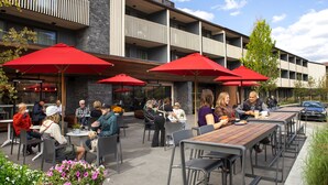 Un área de comedor al aire libre con varias mesas y sombrillas rojas, rodeada por un edificio moderno con balcones.