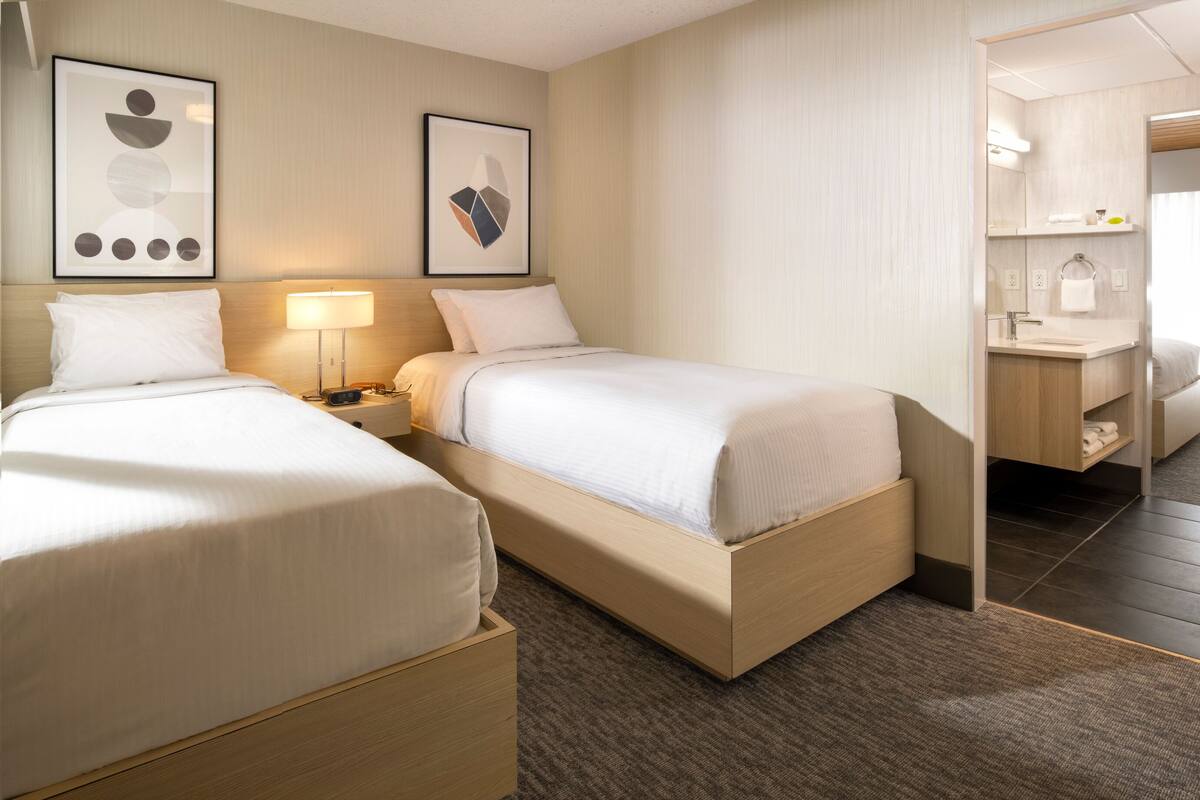 A hotel room with two beds, a desk, a chair, and a bathroom visible through an open door.