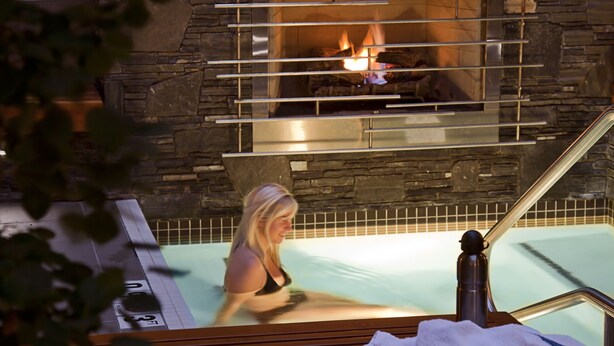 Una chimenea de piedra con fuego, un banco de madera con toallas y una zona de piscina.
