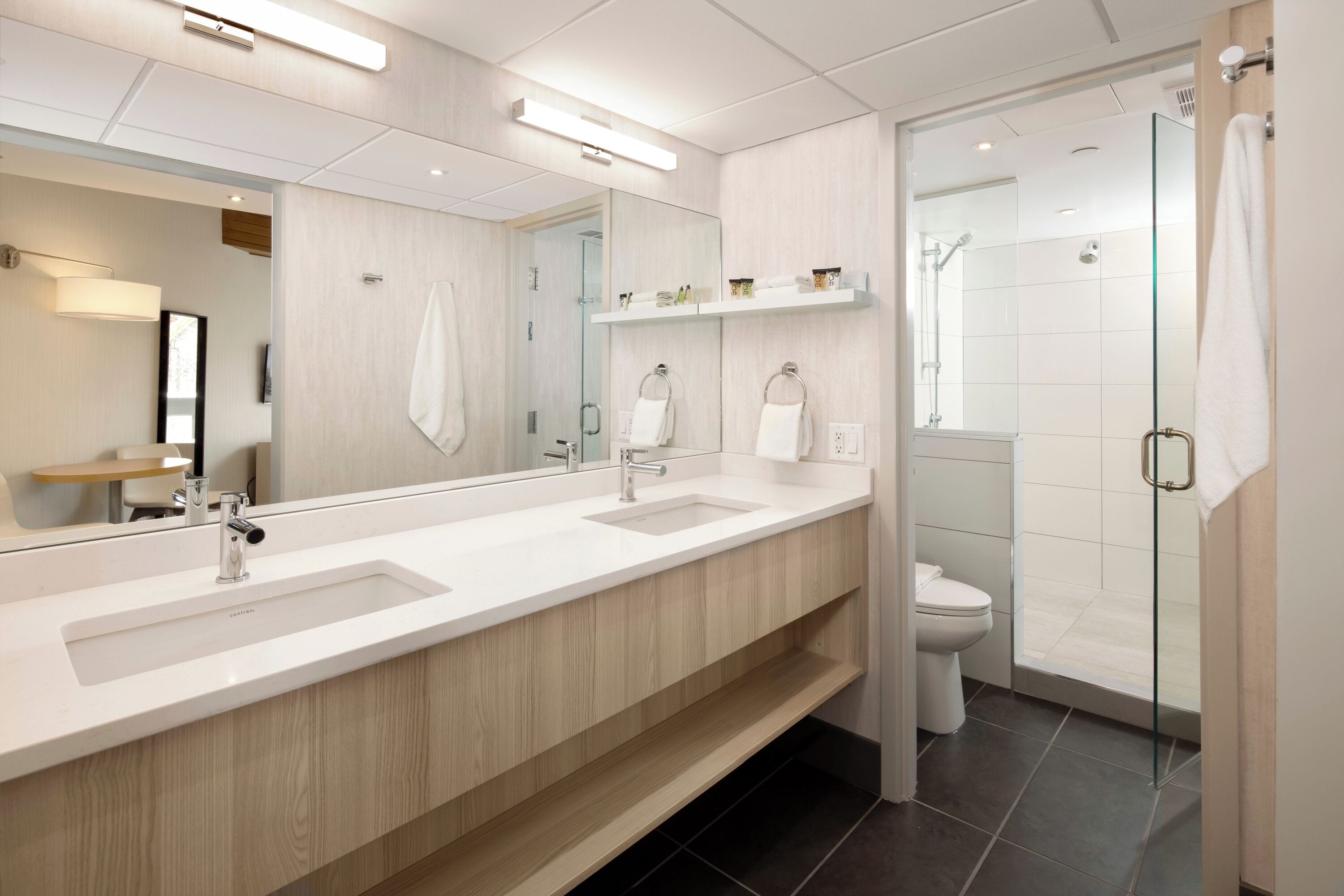 A modern bathroom with a long vanity, two sinks, a large mirror, and a glass-enclosed shower area.
