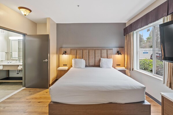 Habitación de hotel con una cama grande, dos mesitas de noche, un cabecero, un televisor, una ventana con cortinas y un baño visible a través de una puerta abierta.