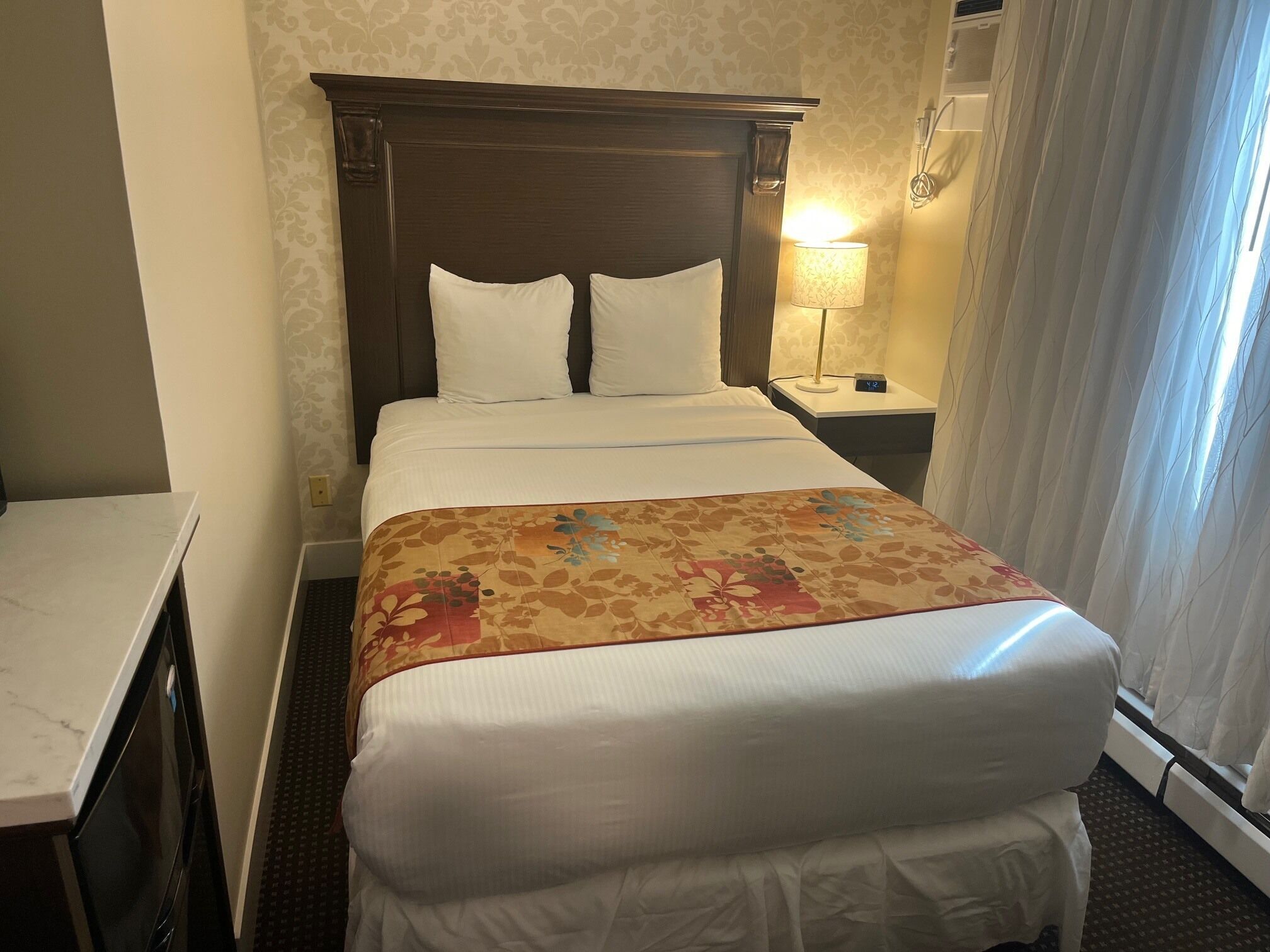 A hotel room with a bed, a nightstand with a lamp, a desk, and a window with curtains.