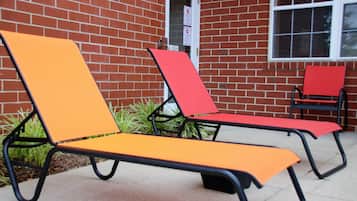 Outdoor lounge chairs with metal frames and fabric seats, positioned against a brick wall with a window.