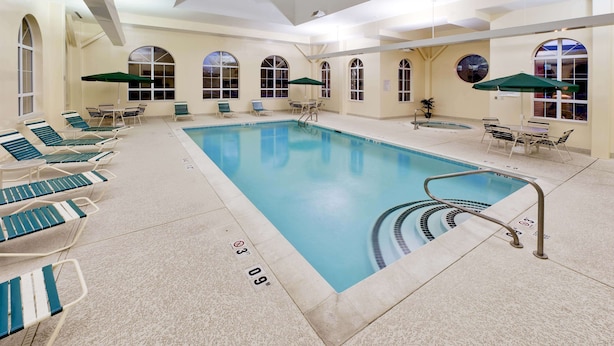 Una piscina interior con sillas de descanso y sombrillas, rodeada de ventanales arqueados y suelo de baldosas.