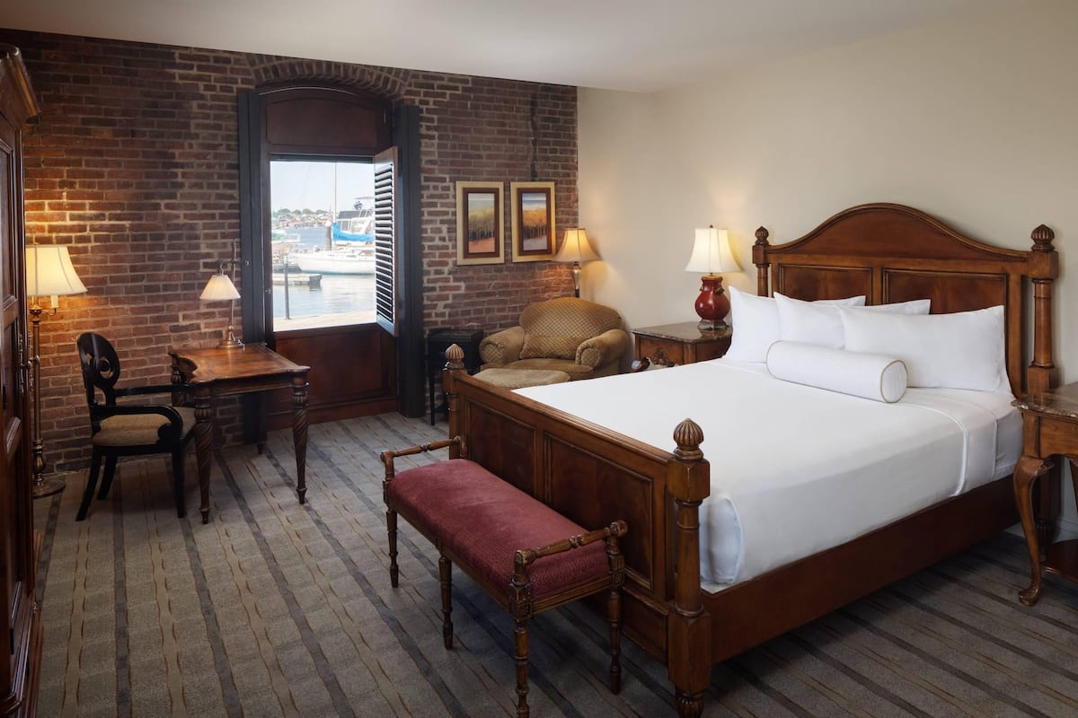 A hotel room with a large bed, a desk, a chair, a bench, and a view of the outside.