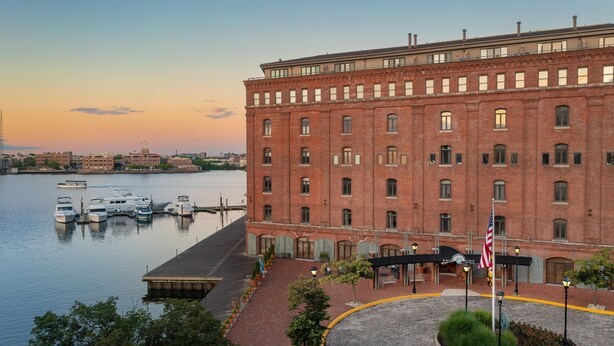 A large brick building with a waterfront view, a flag, and a dock with boats. - The Inn at Henderson's Wharf, an Ascend Collection Hotel (Baltimore)