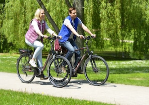 Dos personas andan en bicicleta por un camino pavimentado en un parque con césped verde y árboles.